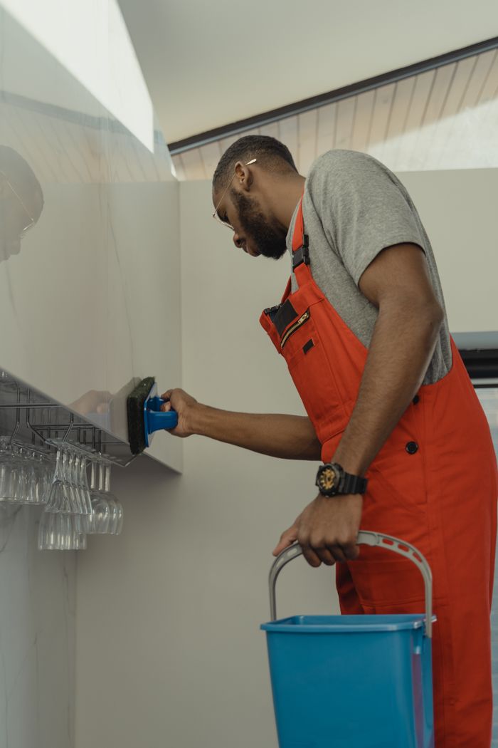 journey Man in red overalls cleaning a glass wall indoors with a blue bucket and brush.